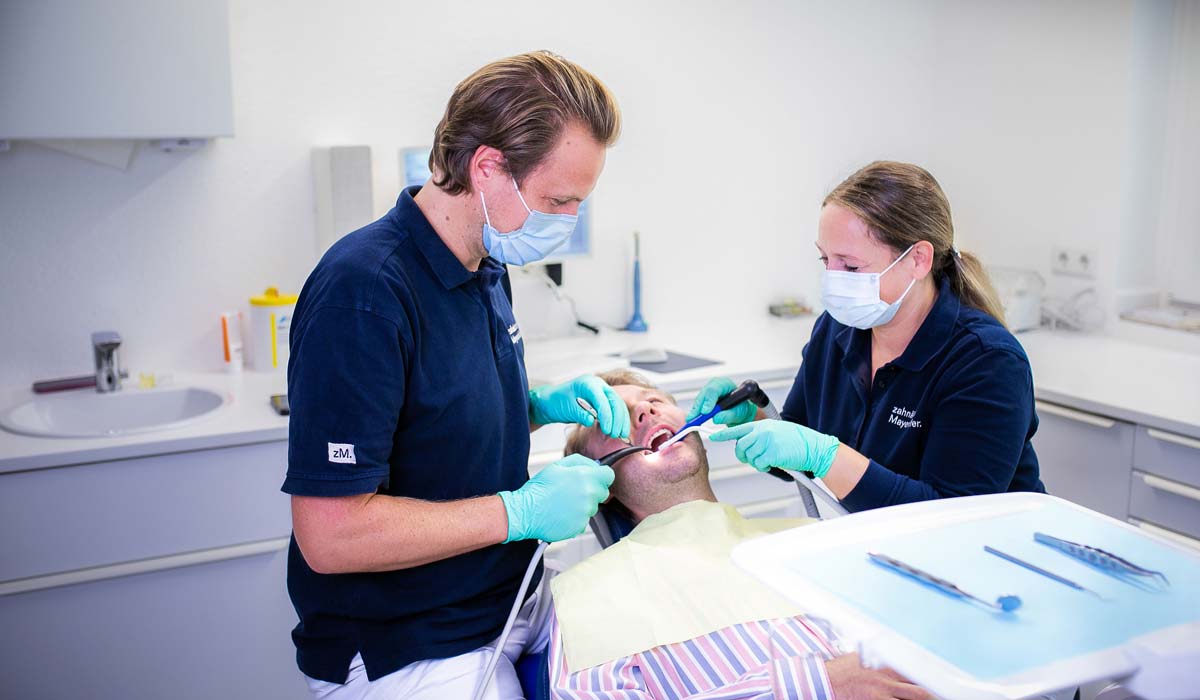 Zahnarzt Dr. David Mayerhöfer behandelt am Behandlungsstuhl in seiner Praxis einen Patienten. Unterstützt wird er durch eine zahnmedizinische Fachangestellte in der Assistenz.